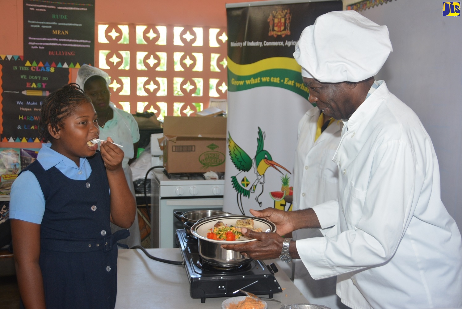 Minister without Portfolio in the Ministry of Industry, Commerce, Agriculture and Fisheries, Hon. J.C. Hutchinson (right), serves breakfast, which he prepared, to student of Holland Primary School in St. Elizabeth, Janessa Clarke, during the Eat Jamaican School Tour at the institution on Friday (November 15).
