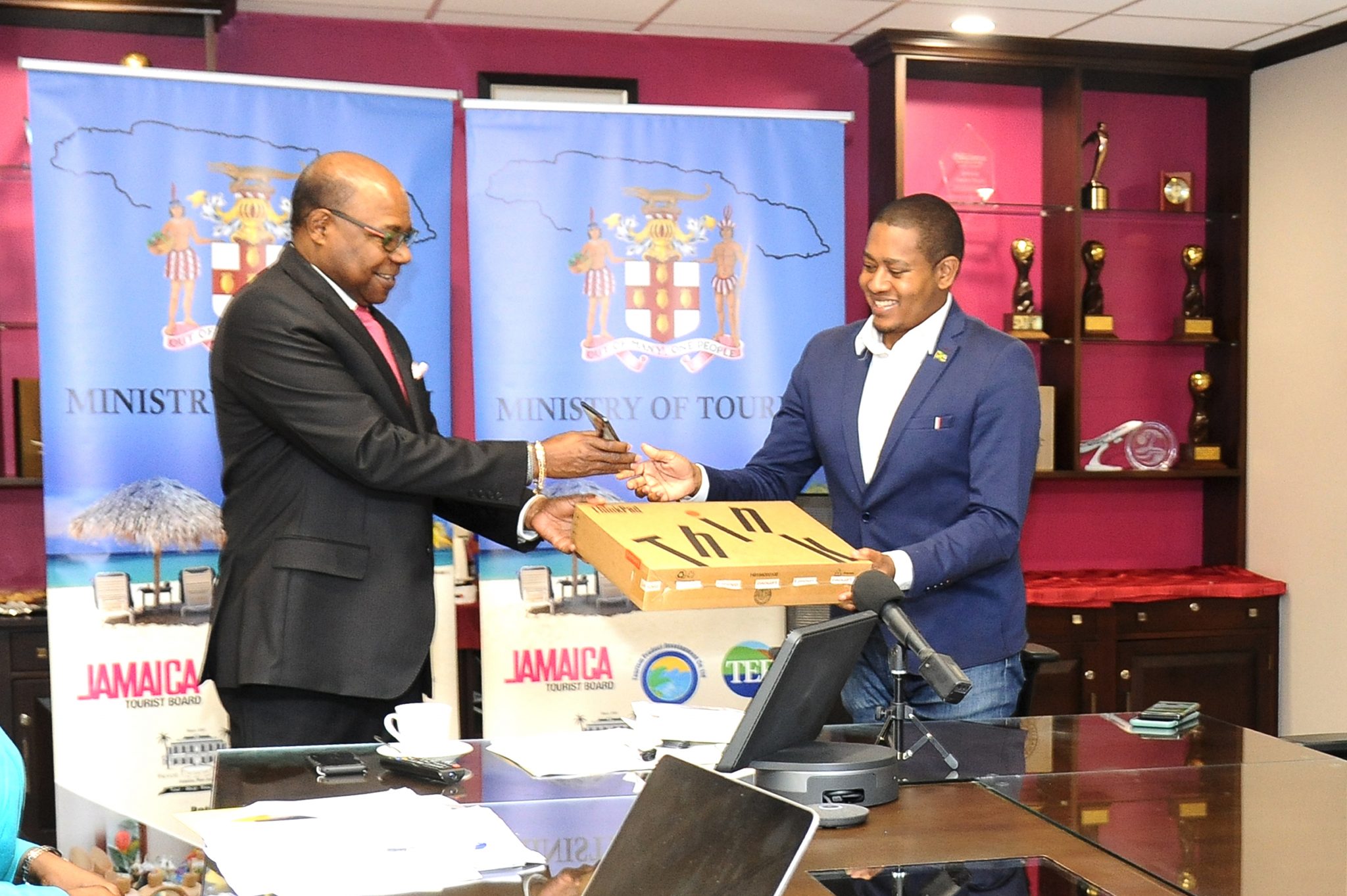 Tourism Minister, Hon. Edmund Bartlett (left), present laptops and  cell phones to Minister of State in the Ministry of Industry, Commerce, Agriculture and Fisheries,  Hon. Floyd Green during a digital press briefing  (April 2) to provide an update on the tourism sector. The equipment, purchased by the Tourism Enhancement Fund at a cost of $1.5 million, will enable staff at the Rural Agricultural Development Authority (RADA) to work from home and have access to the Agri-linkages Exchange (ALEX) platform to help farmers get rid of excess produce.