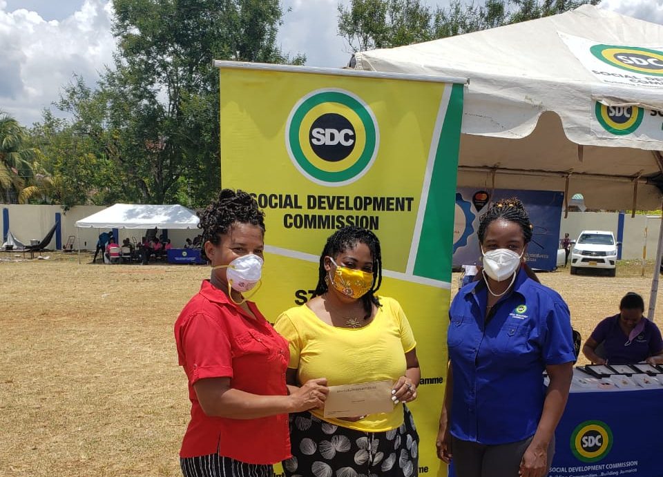 St. Ann Parish Manager for the Social Development Commission (SDC), Hillory McBayne (left); and SDC's Director, Local Economic Development and Community Projects, Avril Ranger (right), present a grant cheque to Melisha Coke of Aisha’s Inspirations, a Local Economic Initiative in St. Ann, at the St. Ann Community Market and Business Fair, held in May 2020.