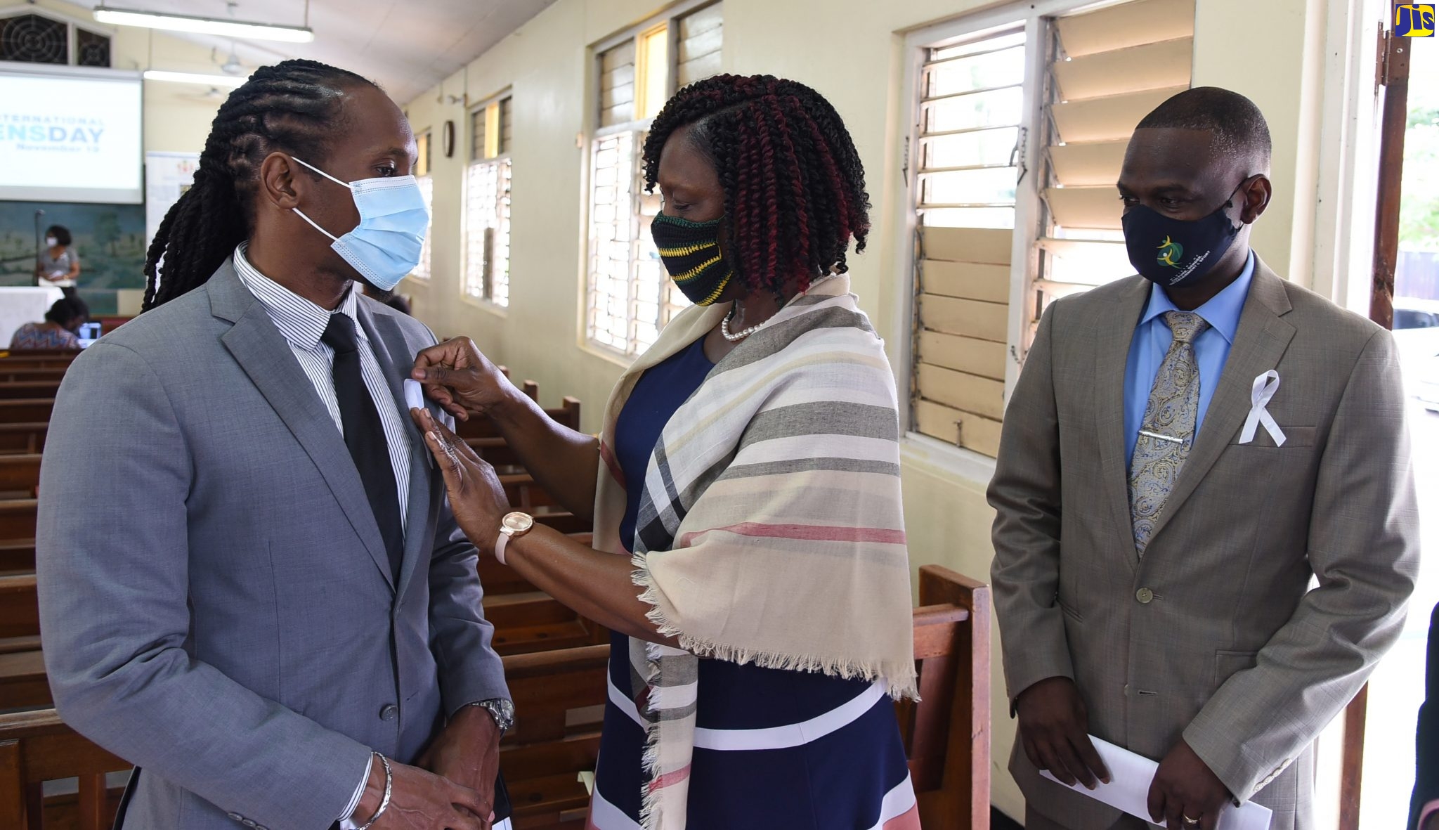 Principal Director, Bureau of Gender Affairs (BGA), Sharon Robinson (centre) pins a white ribbon on the lapel of State Minister in the Ministry of Culture, Gender, Entertainment and Sport, Hon. Alando Terrelonge (left), at the International Men’s Day (IMD) Church Service held on Sunday (November 15) at the Constant Spring Road Church of God, St. Andrew. Looking on is the Permanent Secretary in the Ministry, Denzil Thorpe.
