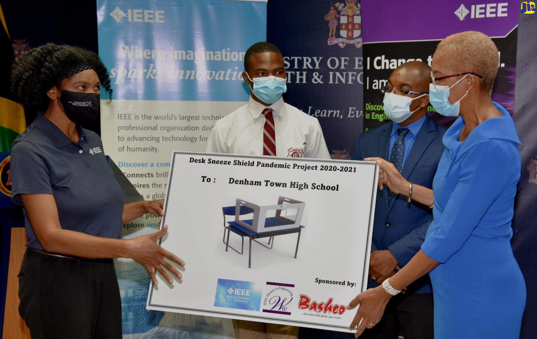 Education, Youth and Information Minister, Hon. Fayval Williams (right), and Chair, Institute of Electrical and Electronics Engineers (IEEE) Jamaica Women in Engineering Affinity Group, Charlene Brown (left), display a photo of one of 350 desk shields being donated by the organisation to the Denham Town High School in Kingston. The occasion was a symbolic handover ceremony for the shields at the Ministry’s offices in Kingston on Friday (June 25). Looking on are Denham Town High student, Taquan Graham (2nd left), and Acting Principal, Donovan Hunter. 