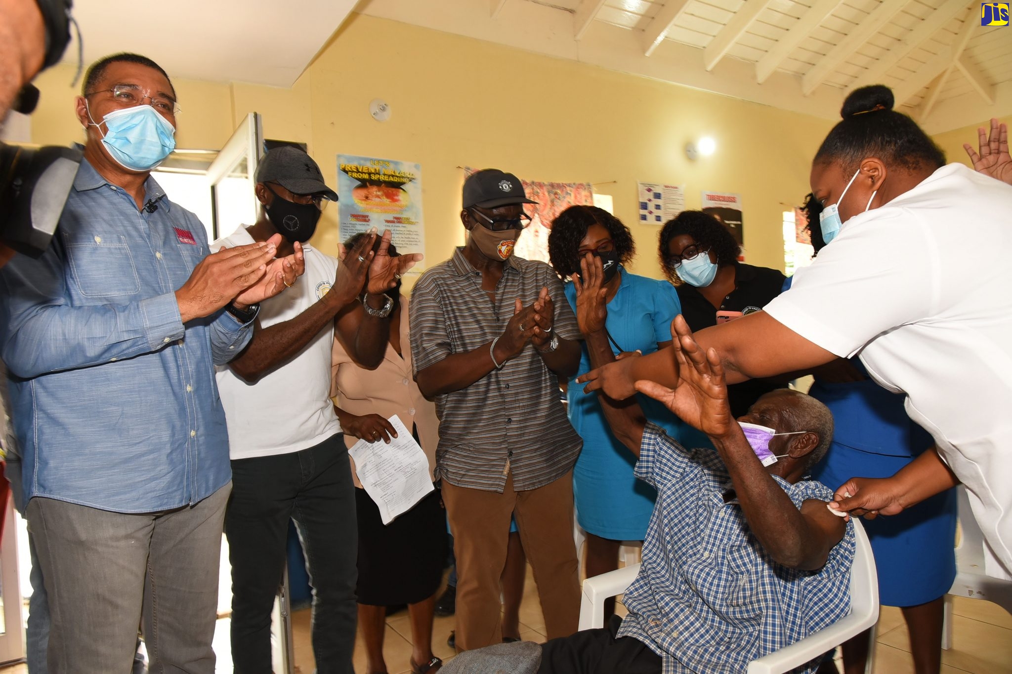 Prime Minister, the Most Hon. Andrew Holness (left), applauds as farmer, Leslie Sharpe  (seated-right), takes the coronavirus (COVID-19) vaccine, being administered by nurse Sasha Ford (right) at the James Hill Health Centre in Clarendon, recently. Sharing in the moment (from left) are Member of Parliament for Clarendon Northern, Dwight Sibblies; Minister of Local Government and Rural Development, Hon. Desmond McKenzie; and other healthcare professionals.