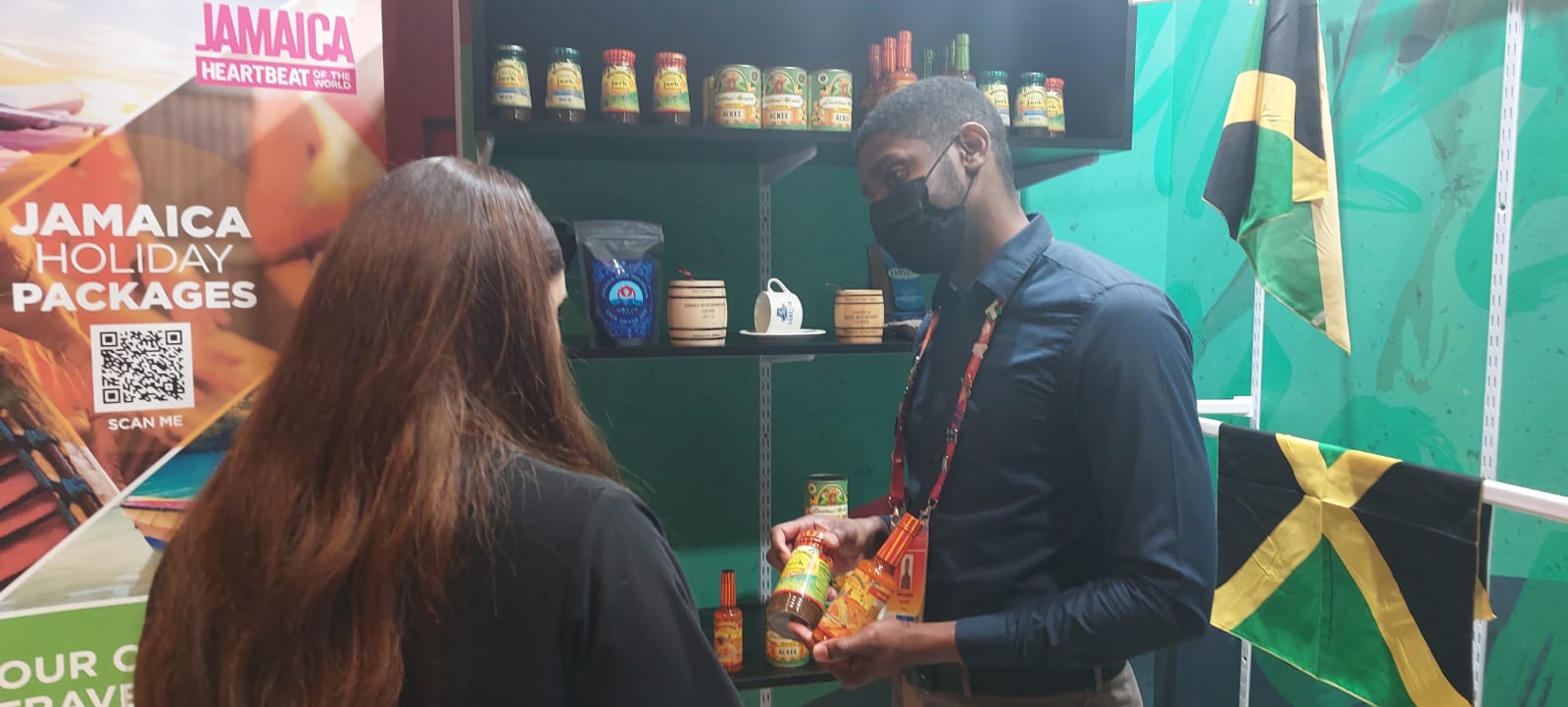 Manager, Business Facilitation and Acceleration Services at the Jamaica Special Economic Zone Authority (JSEZA), Kevon Farquharson (right). explains the difference in flavour profiles and uses between a Walkerswood  jerk marinade and a Walkerswood pepper sauce, to a visitor of the Jamaica Pavilion at World Expo 2020 Dubai. The Walkerswood products are among several Jamaican products available for purchase inside the Pavilion. Mr. Farquharson says  the rate at which Jamaican products are being purchased from the Pavilion is an indicator of the demand in the area and is a market for the country to get into. 