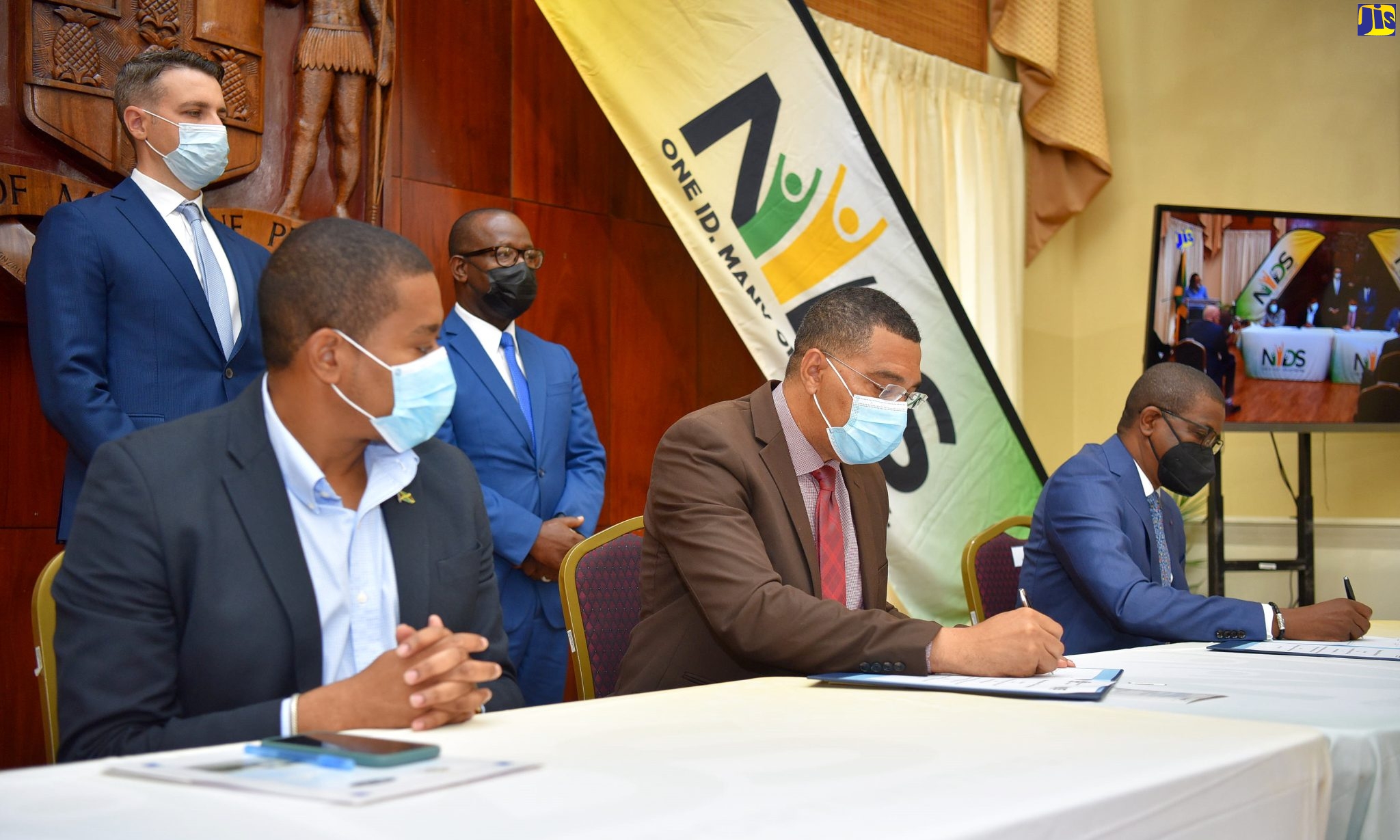 Prime Minister, the Most Hon. Andrew Holness (seated centre), and President, Jamaica Bankers Association and Chief Executive Officer, National Commercial Bank (NCB), Septimus Blake (seated left), affix their signatures to the Memorandum of Understanding (MOU) with deposit-taking institutions for the National Identification System (NIDS) pilot. The signing ceremony was held at the Office of the Prime Minister in St. Andrew on June 3. Observing the signing are Minister without Portfolio in the Office of the Prime Minister, Hon. Floyd Green (seated left); Modernisation of State Specialist, Inter-American Development Bank, Benjamin Roseth (standing, left) and NIDS Programme Director, Warren Vernon.