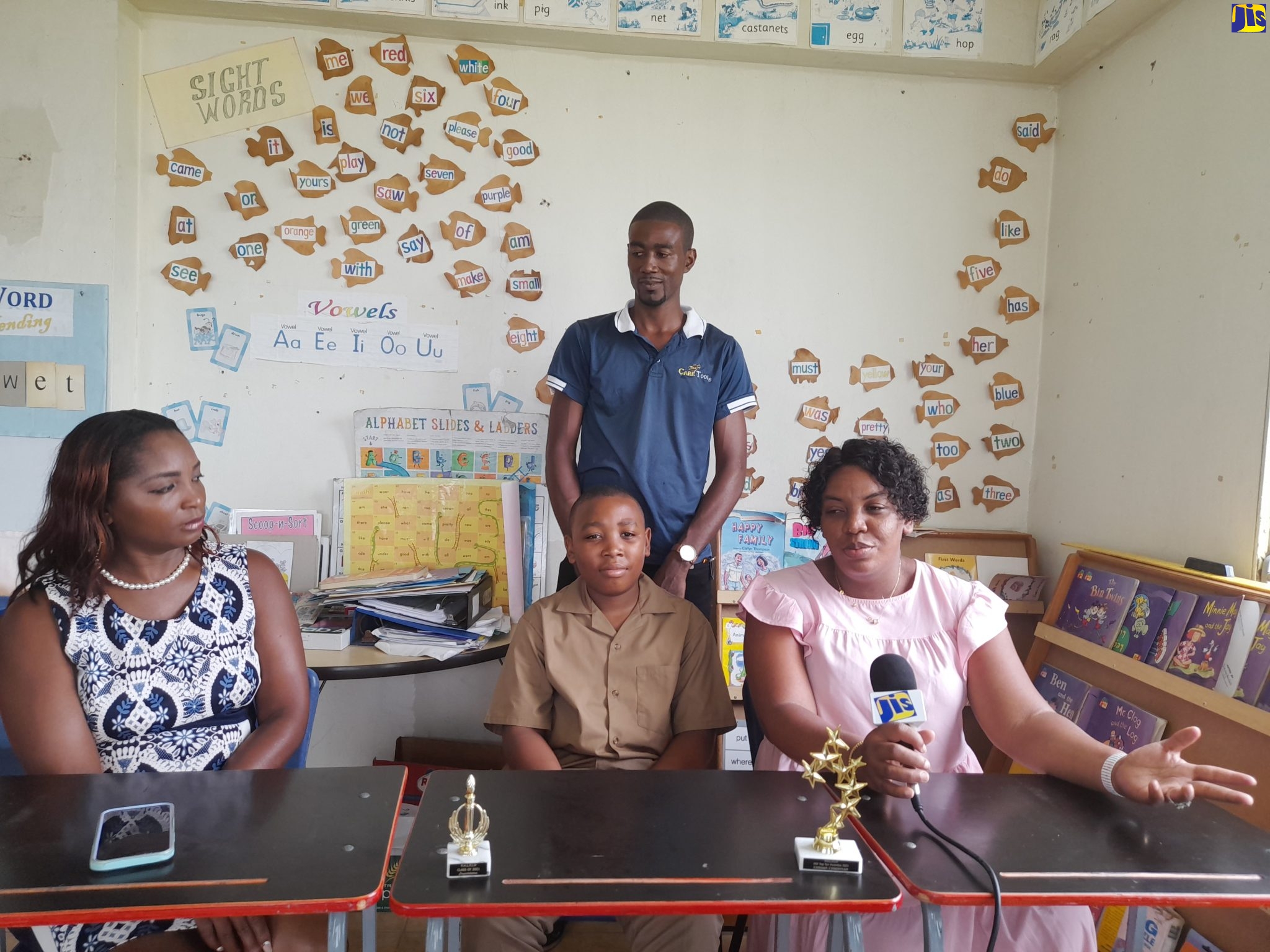 Principal of the Point Hill Leased Primary School in St. Catherine, Shernette Powell (left), looks on while early-childhood educator and parent of the top-performing student at the Point Hill primary, Nadine Sinclair Peart (right), makes a point, during a recent interview with JIS News. Others pictured are top-student Naveed Peart (seated), and his father, James Peart. 