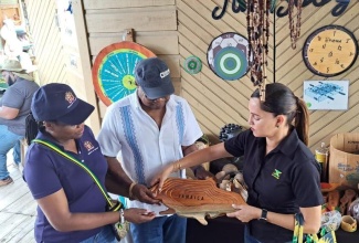 Minister of Tourism, Hon. Edmund Bartlett (centre); Permanent Secretary in the Ministry, Jennifer Griffith (left) and Member of Parliament for Eastern Portland, Annmarie Vaz, look at a carving in the shape of the island of Jamaica, during a recent stop at the Port Antonio Craft Market, during a Destination Assurance Tour of sections of Portland.