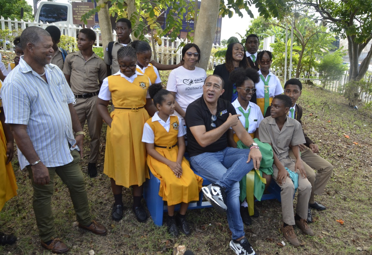 Minister of Health and Wellness, Dr. the Hon. Christopher Tufton (seated, second left), sits on a wellness bench with students of the B.B. Coke High School in St. Elizabeth. The bench was unveiled during a mental wellness session held at the school on Thursday (October 26) in collaboration with the Ministry of Education and Youth. Joining the Minister are Chairman of the school board, Cetany Holness (left); Acting Principal of B.B. Coke High, Liteasha Gallimore (standing, third right); and Director of Child and Adolescent Mental Health in the Ministry of Health and Wellness, Dr. Judith Leiba (standing, fourth right).