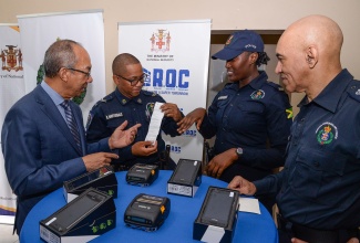 Deputy Prime Minister and National Security Minister, Hon. Dr. Horace Chang, observes as Jamaica Constabulary Force (JCF) Officer, Byron Martindale, exhibits a ticket generated by the e-ticketing system during a demonstration of the handset device by Constable Morata Murdock. Also observing is Police Commissioner, Major General Antony Anderson. The occasion was the handover of 3,000 handsets and printers to the JCF’s Public Safety Traffic Enforcement Branch (PSTEB) in Kingston in December.

