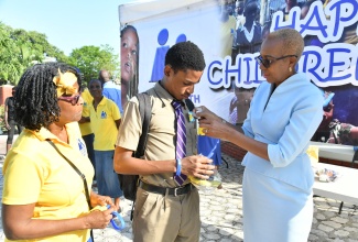 Minister of Education and Youth, Hon. Fayval Williams (right), places a sticker on Kingston College student, Ricquan Bailey, as part of activities marking Children’s Day at St. William Grant Park in downtown Kingston on Friday (May 17). The event was organised by the National Child Month Committee (NCMC). Looking on is NCMC Chair Emeritus, Dr. Pauline Mullings.

