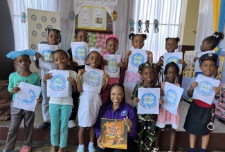 Students from St. Joseph’s Infant School in Kingston display  the National Land Agency's logo during Read Across Jamaica Day on May 7.  Seated is the  Agency’s Manager of Marketing and Public Relations, Nicole Hayles, who read to the students.

