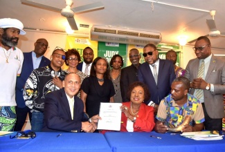 Minister of Culture, Gender, Entertainment and Sport, Hon. Olivia Grange, and Group Head of Life, Health and Pensions at Guardian Life Insurance, Eric Hosin, display the signed contract for the Jamaica Entertainers and Creatives Insurance Plan (JECIP). They are surrounded by stakeholders, including entertainers and creatives. The signing ceremony took place on Tuesday (November 12) at the Ministry of Culture, Gender, Entertainment and Sport in Kingston.