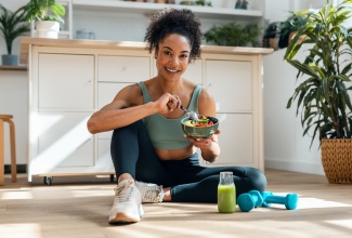 Picture of woman in athletic gear eating a fruit bowl while sitting on floor in the kitchen at home.