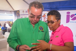 Minister of Science, Energy, Telecommunications and Transport, Hon. Daryl Vaz and Customer Service Representative at Jamaica Public Service Company (JPS), Rashida Leiba, look at the power company’s app, during the launch of the Government’s Community Electrification Project held at the Rhules Pen (Dampy) Playfield in Mineral Heights, Clarendon.