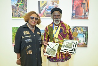 Minister of Culture, Gender, Entertainment and Sport, Hon. Olivia Grange (left), shares a photo moment with Most Outstanding Overall Artist, Nugent Hewitt, 72, who received six gold medals and one silver medal in the 2025 Jamaica Cultural Development Commission (JCDC) Visual Arts Competition. Displayed behind them are some of his award-winning pieces featured in the Gold Medal Winners Exhibition, held recently at the Olympia Art Centre on Old Hope Road in Kingston.

