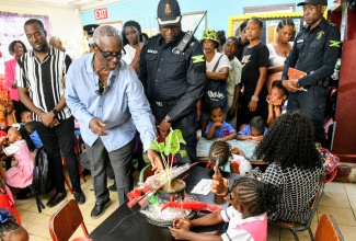 Minister of Local Government and Community Development, and Member of Parliament for West Kingston, Hon. Desmond McKenzie (centre), is flanked by Senior Superintendent of Police (SSP) for the Jamaica Constabulary Force (JCF) Area 4 Headquarters, Michael Bailey (right), and Councillor for the Denham Town Division, Dellon Gayle (left), as they view the memorial set up on Jayce Pinnock’s desk at the Edward Seaga Infant School in Denham Town, Kingston, during a visit on Monday (February 23). Jayce was fatally shot and his father and sibling injured in a gun attack at their home in Denham Town on Saturday (February 21).