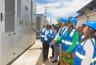 Minister of Health and Wellness, Dr. the Hon. Christopher Tufton (third right), is joined by National Health Fund (NHF) Board Chairman Shane Dalling (second left); NHF Chief Executive Officer, Everton Anderson (third left); Speaker of the House of Representatives, the Most Hon. Juliet Holness (second right); and Vice President of Programme Operations at Direct Relief, Genevieve Bitter (right), along with other stakeholders, as they listen to NHF Director of Institutional Benefits, Projects and Maintenance, Richard Allen (left), explain components of the solar system installed at the NHF