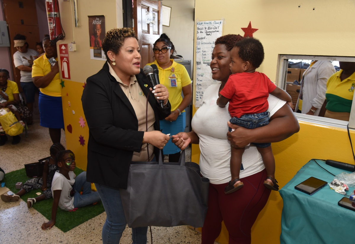 Minister of State in the Ministry of Health and Wellness, Hon. Krystal Lee (left), presents a gift package to mother Shaquan Nelson, who is holding her daughter, Rasheeda Henry, during a visit to the Glen Vincent Health Centre in St. Andrew on Friday (May 15).
