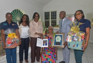 Minister of Labour and Social Security, Hon. Pearnel Charles Jr. (second right), presents 100-year-old Dr. Mavis Gilmour Petersen (seated third right) with a plaque recognising her as an outstanding centenarian with a legacy of service, during a visit to her home in Cherry Gardens, St. Andrew, on Wednesday (May 20), in observance of Centenarians Day 2026. Sharing the moment (from left) are Parish Organiser, National Council for Senior Citizens (NCSC), Shonelle Lewis-Mckie; Executive Director of the NCSC, Cassandra Morrison; Acting Permanent Secretary in the Ministry, Dione Jennings; and Programme Organiser, NCSC, Marieck Hendricks-Morgan.