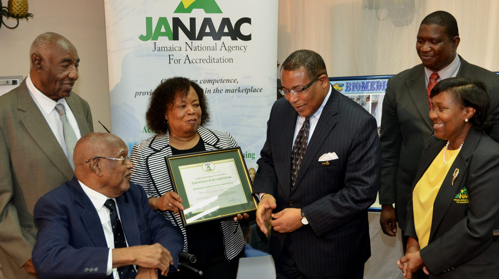 Minister of Industry, Investment and Commerce, Hon. Anthony Hylton (3rd right) along with Chief Executive Officer of the Jamaica National Agency for Accreditation (JANAAC), Marguerite Domville (right) ; and JANAAC Chairman, Dr. Cliff Riley (2nd right),  congratulate Chairman of the Board of Biomedical Caledonia Medical Laboratory, Dr. Donald Christian (seated), after presenting an accreditation certificate from JANAAC. Holding the certificate is Managing Director of Biomedical, Barbara Hendriks, and  Board member, Lester Woolery is at left. The presentation  ceremony was held at Hotel Four Seasons, today (June 25).
