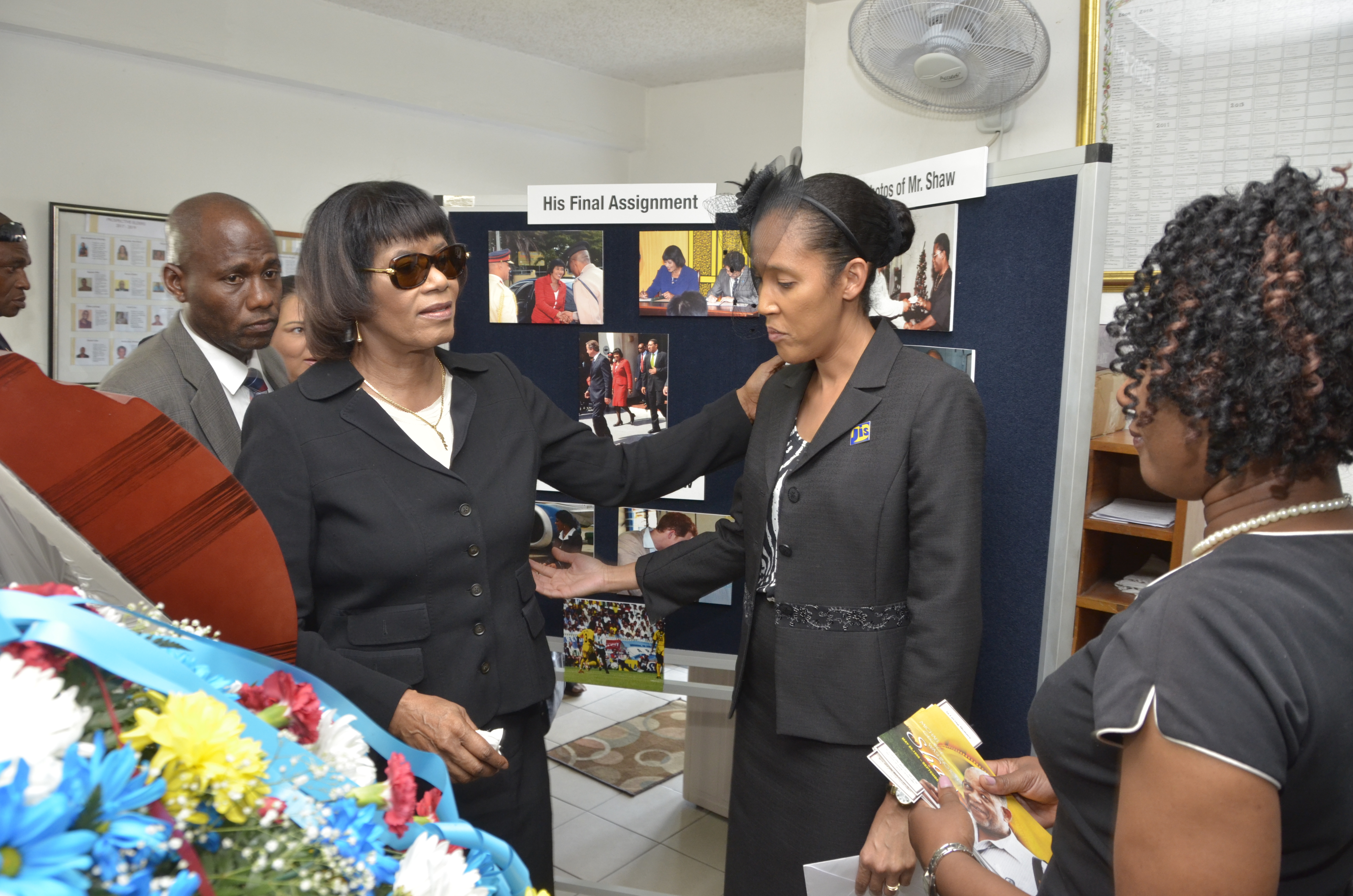 Prime Minister, the Most Hon. Portia Simpson Miller (2nd left), consoles Chief Executive Officer of the Jamaica Information Service (JIS), Donna Marie Rowe (2nd right), just before the start of the thanksgiving service for late veteran photographer, Michael Shaw, on October 23,  at the Webster Memorial United Church in Kingston. In the background is an exhibit of Mr. Shaw’s work over the years, including pictures from his final assignment on September 30.
