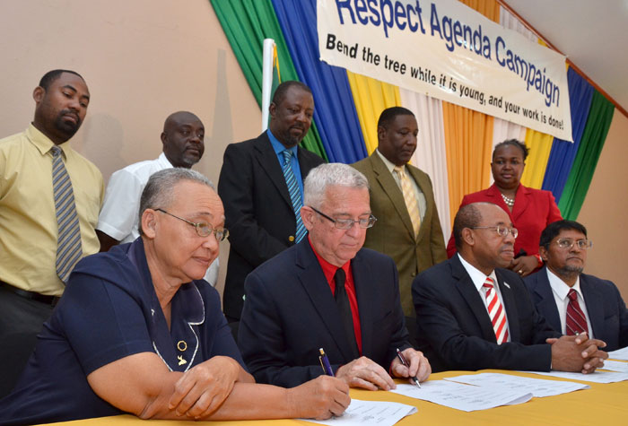 Minister of Education, Hon. Rev. Ronald Thwaites (seated, second left); and Chairman of the Portmore Cluster of Schools and Principal of the Portmore HEART Academy, Karen Kennedy; affix their signature to a Memorandum of Understanding which solidifies the collaboration of a cluster of Portmore-based schools in advancing the Ministry’s ‘Respect Agenda’ campaign, while  Executive Director, PetroCaribe Development Fund, Dr. Wesley Hughes (second right); and Campaign Programme Co-ordinator, Reginald Budhan (right); look on. Other signatories pictured (standing, from left), are Principals of Waterford High, Oliver Holness; Ascot High, Cedrick Murray; Bridgeport High, Aston Messam; and Cumberland High, Michael Brydson while Chief Education Officer, Grace McLean is at right. The signing took place at the Portmore HEART Academy in St. Catherine on February 14.