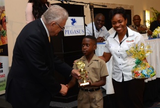 Minister of Industry, Commerce, Agriculture and Fisheries, Hon. Karl Samuda, presents the Ministry’s trophy to Asher Harrison of Greater Portmore Primary School, who was the winner in the primary school category of the Praedial Larceny Prevention Unit (PLPU) inaugural essay competition in 2016. Sharing the moment is Marketing Coordinator at Newport Fersan, Joan Sharpe Colley.