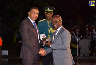 Prime Minister, the Most Hon. Andrew Holness (left), presents Fabian Brown (right) with his award for Community service and Development. Mr. Brown was among 65 persons who were conferred with the Prime Minister’s Jamaica 55 Commemorative Medal of Appreciation for 2017, during a ceremony held on the lawns of Jamaica House on Wednesday (April 25). Assisting is aide-de-camp assigned to the Prime Minister for the function, Major Karl McKen.

