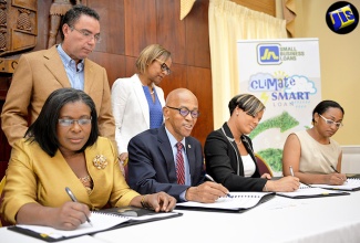 Minister without Portfolio in the Ministry of Economic Growth and Job Creation, Hon. Daryl Vaz (standing, left) and General Manager, Country Department, Caribbean Group, Inter-American Development Bank, Therese Turner Jones (standing, right), observe as (from left) Permanent Secretary in the Ministry of Economic Growth and Job Creation, Audrey Sewell; Managing Director, Development Bank of Jamaica, Milverton Reynolds; General Manager, JN Small Business Loans, Gillian Hyde; and Programme Manager, Environmental Foundation of Jamaica (EFJ), Allison Rangolan McFarlane sign the Memorandum of Understanding (MOU) for the Adaptation Programme and Financing Mechanism Project at Jamaica House, in St. Andrew, on July 28. Under the initiative, US$7.2 million will be made available to micro, small and medium-size enterprises (MSMEs) in the tourism and agricultural sectors to finance climate-change adaptation initiatives islandwide.  