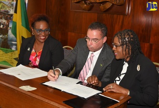 Minister without portfolio in the Ministry of Economic Growth and Job Creation, Hon. Daryl Vaz (centre), affixes his signature to the agreement for the provision of a US$300,000 grant from the United Nations (UN) Green Climate Fund (GCF) to strengthen the country’s capacity to respond to climate change. Looking on (from left) are Principal Director in the Climate Change Division, Una May Gordon; and Permanent Secretary in the Office of the Prime Minister (OPM), Audrey Sewell. The signing ceremony was held on Tuesday (October 17) at the  OPM in St. Andrew.