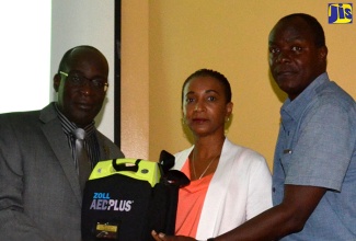 Minister of Education, Youth and Information, Senator the Hon. Ruel Reid (left) and parents of the late schoolboy footballer, Dominic James, Denese (centre) and David James (right), display one of the 15 Automated External Defibrillators (AEDs), presented to schools across the island by Team Jamaica Bickle, at a presentation ceremony held today, (October 14), at JAMPRO head office, in New Kingston. 