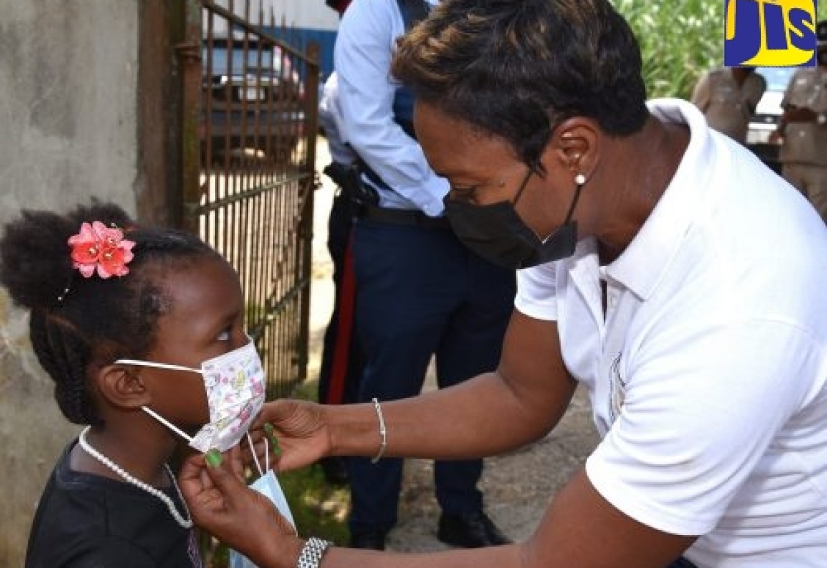 Minister of State in the Ministry of Health and Wellness and Member of Parliament for St. Andrew West Rural, Hon. Juliet Cuthbert Flynn (right), assists young Melissa Samuels in putting on her mask during a recent tour of communities in the constituency.