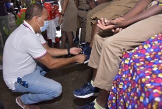 Minister of Health and Wellness, Dr. the Hon. Christopher Tufton (left), ties the shoe lace of a student at Holy Trinity High School, who received sneakers through a donation made possible by the non-profit organisation, Sole 2 Sole.  This was during a ceremony held at the School located on George Headley Drive, in Kingston on February 4.