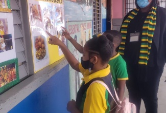 Grade four students of Corinaldi Avenue Primary School in St. James Jhiana Sinclair (left) and Marquin Thorpe (centre) read a culinary-themed display at the school during Jamaica Day on Friday, February 25. Looking on is Acting Principal of the institution, Deon Stern-Anglin.