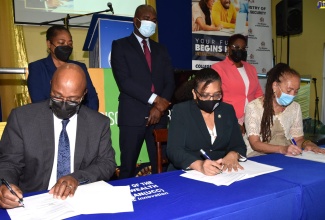 State Minister in the Ministry of National Security, Hon. Zavia Mayne (standing centre), observes the signing of a Memorandum of Understanding (MOU) to facilitate the award of six full tuition scholarships for four inmates, and two correctional services officers to study for an Associate Degree in Business Administration at the University of the Commonwealth Caribbean (UCC). The Signatories are (seated from left) Commissioner of Corrections, Department of Correctional Services (DCS), Lieutenant Colonel (Ret.), Gary Rowe; Deputy Executive Chair, University of the Commonwealth Caribbean (UCC) Group, Geraldine Adams; and Executive Director, Stand up for Jamaica, Carla Gullotta. Also witnessing the proceedings are Head of Department, Business Administration UCC, Sonia Davidson (standing left); and Chief Technical Director in the Ministry, Shauna Trowers. The signing took place on Wednesday (February 23) at the UCC in  Kingston, and livestreamed on the DCS’ Instagram page.