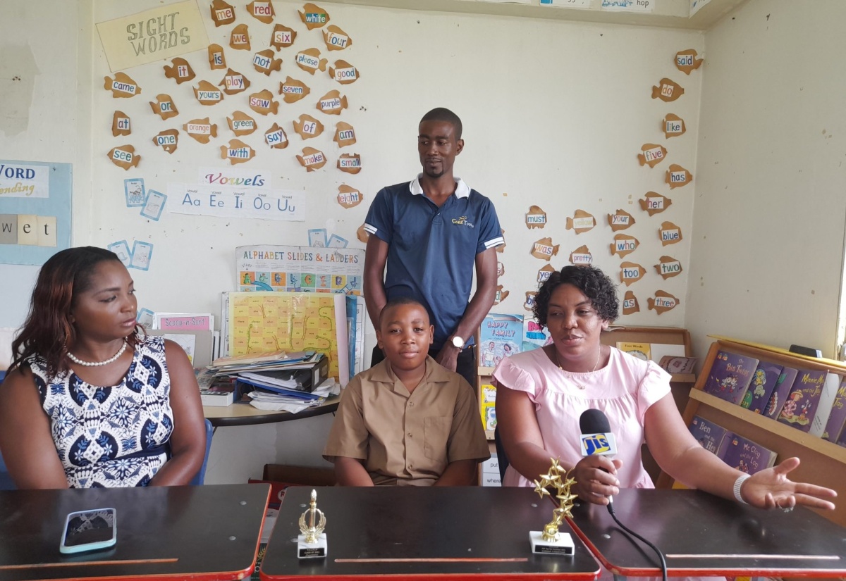 Principal of the Point Hill Leased Primary School in St. Catherine, Shernette Powell (left), looks on while early-childhood educator and parent of the top-performing student at the Point Hill primary, Nadine Sinclair Peart (right), makes a point, during a recent interview with JIS News. Others pictured are top-student Naveed Peart (seated), and his father, James Peart. 