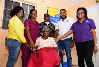 Pauline Forbes (seated) is all smiles as she sits in the new wheelchair she received from the Registrar General’s Department (RGD) and Food For the Poor Jamaica, today (December 20). Surrounding her are (from left) RGD Parish Officer for Kingston, Tamar Blake; Geriatric Nurse, Georgia Currie-Taylor; her husband, Hubert Forbes; Chief Executive Officer of the RGD, Charlton McFarlane and Marketing and Planning Manager at the RGD, Nicole Whyte. 