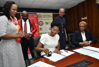 Minister of Education and Youth, Hon. Fayval Williams (background right), looks on as Senior Operations Manager, Digicel Foundation, Jodi-Ann McFarlane (seated left) stamps the document formalising the partnership between the entities for the renovation of the Santa Cruz Basic School to establish the Santa Cruz Special Education Centre. The signing ceremony was held at the Ministry’s National Heroes Circle offices in Kingston on January 11.  Observing (from left) are Acting Chief Education Officer in the Ministry, Dr. Kasan Troupe; Director of Regional Education Services at the Ministry, Region 5, Ottis Brown and Acting Permanent Secretary in the Ministry, Maureen Dwyer.