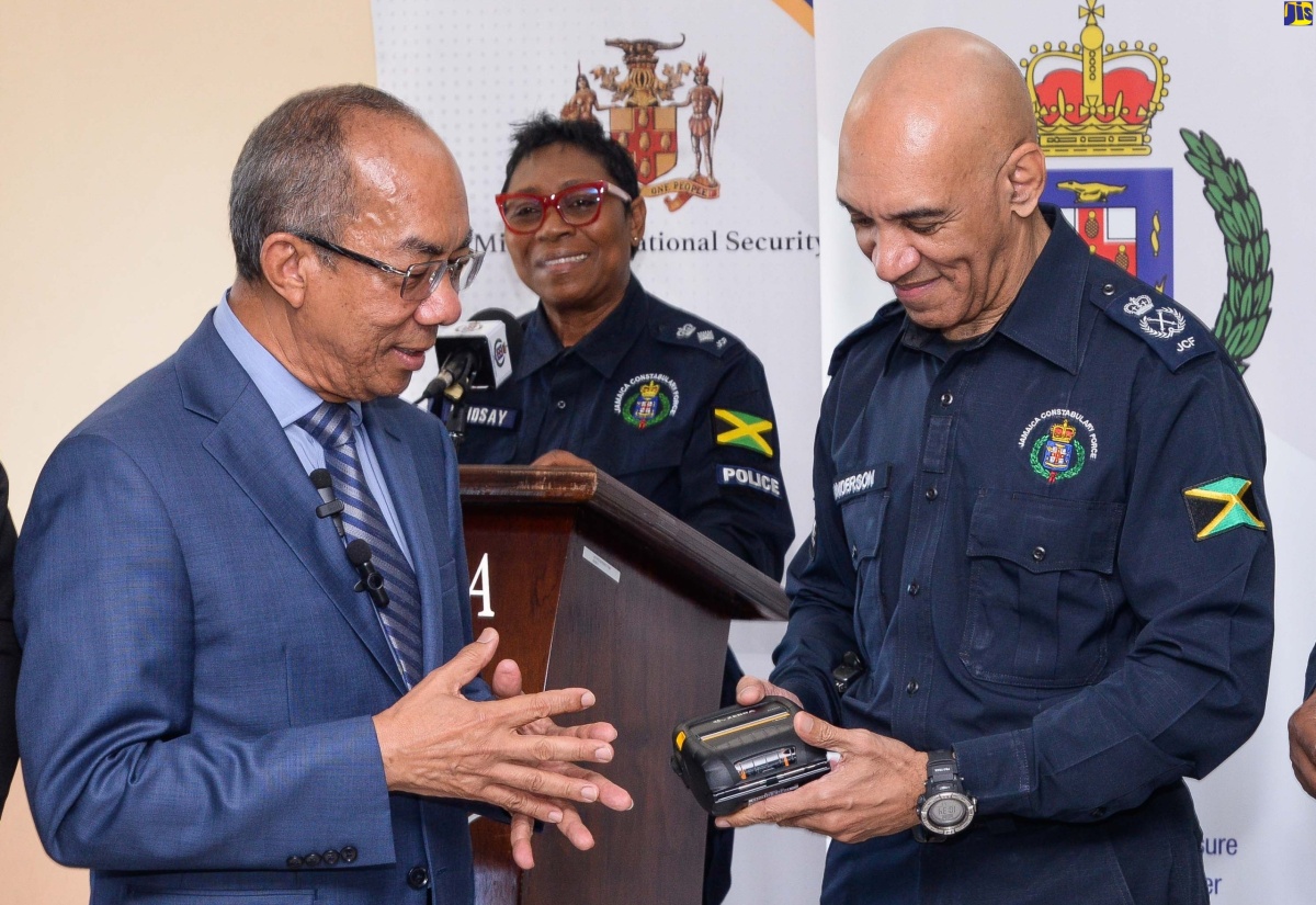 National Security Minister, Hon. Dr. Horace Chang (left), and Commissioner of Police, Major General Antony Anderson, look at an e-ticket printer, during the handover of 3,000 sets of rugged handsets and printers at Public Safety and Traffic Enforcement Branch (PSTEB) on Elletson Road in Kingston on December 19. At the podium (centre, background) is Senior Superintendent of Police, Stephanie Lindsay.