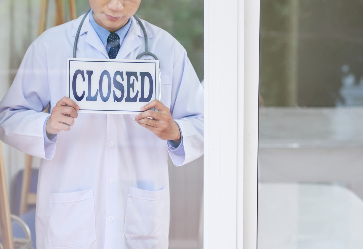 Man in medical uniform holding sign with 'Closed' writing while standing behind glass in office.