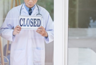 Man in medical uniform holding sign with 'Closed' writing while standing behind glass in office.