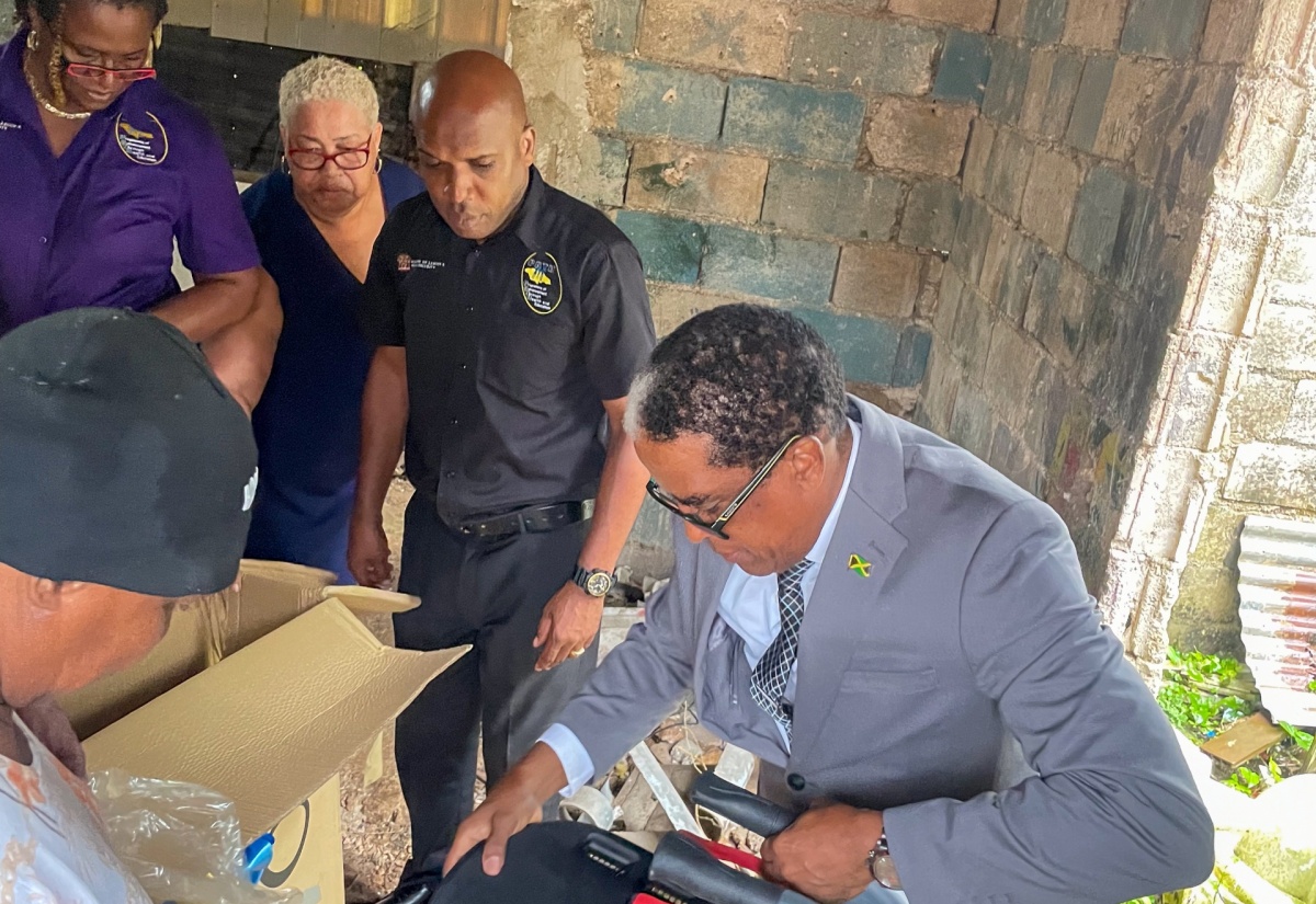 State Minster in the Ministry of Labour and Social Security, Dr. the Hon. Norman Dunn (right), assembles the Rollator Walker, during the handover of items to Murinne Steer (left) for her disabled son, at Liberty Valley in Brown’s Town, St. Ann, on February 20.  Others looking on (from second left) are Divisional Director for Social Security in the Ministry, Suzette Morris; Corporate Communications Manager, Marcia Williams; and Parish Administrator for the Ministry’s St. Ann office, Ansel Rob.


