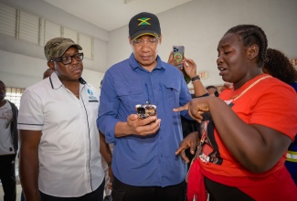 Prime Minister, the Most Hon. Andrew Holness (centre), looks at cell phone images of damage to housing being pointed out by Rocky Point resident, Rochelle “Shanice” Brooks, during a visit to the Southern Clarendon community on Saturday (July 6). He was accompanied by Minister of Labour and Social Security, the Hon. Pearnel Charles Jr. who is also the Member of Parliament for Clarendon Southeast.