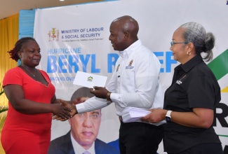 Minister of Labour and Social Security, Hon. Pearnel Charles Jr., presents a cheque to Belinda Bowen, during the handover of cash grants to persons across the parish of St. Catherine whose houses were damaged by Hurricane Beryl. The ceremony was held on August 27, at the HEART College of Construction Services in Portmore. At right is Permanent Secretary in the Ministry, Colette Roberts Risden.

